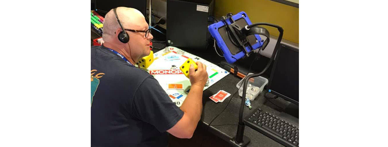 elder white man plays monopoly - he is wearing headphones and is looking at an ipad on an eye level stand, angled to see the board by a second virtual player