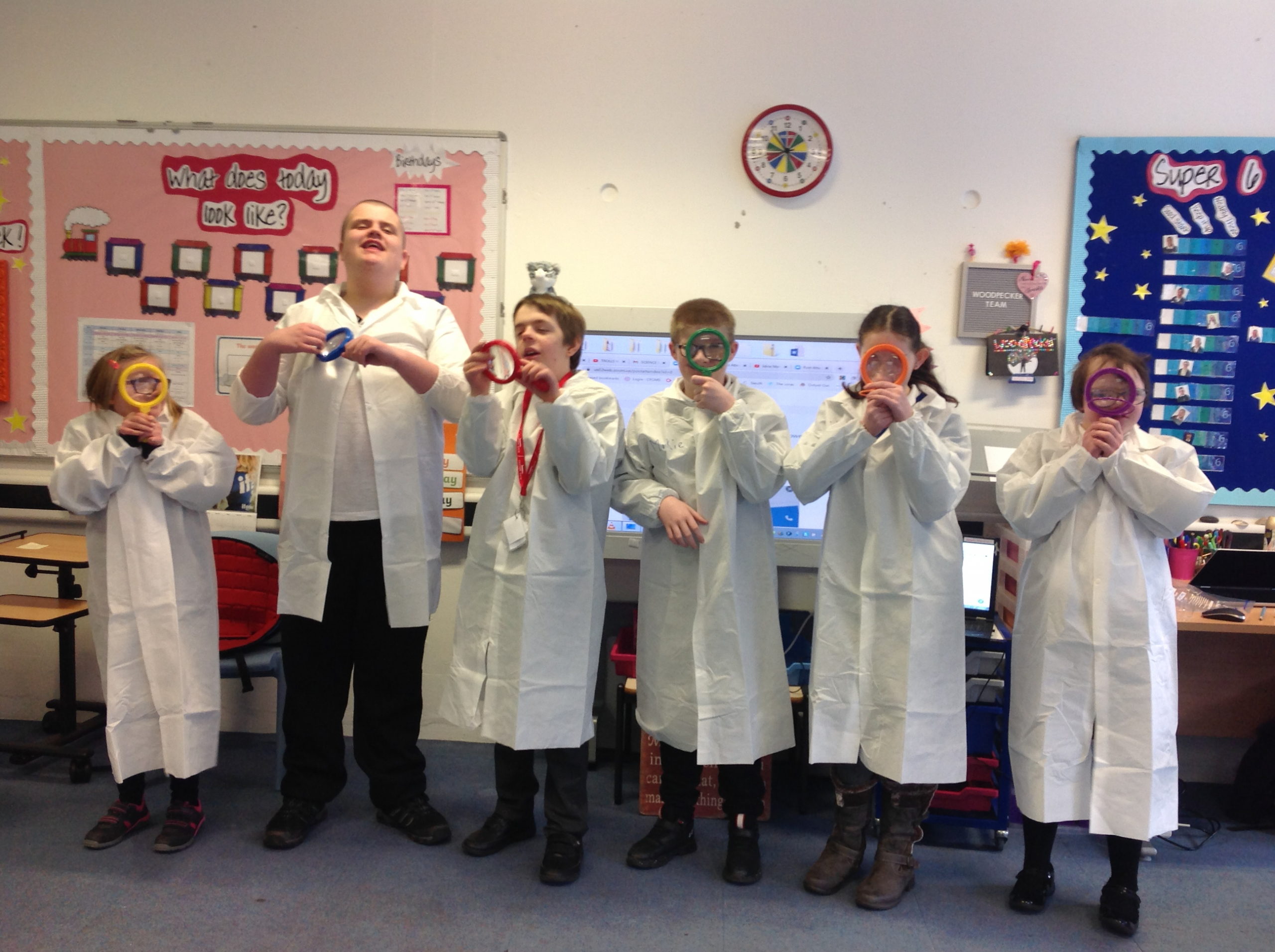 Six young people with learning disabilities stand in a line. They are in a classroom and are all wearing white lab coats. They have large magnifying glasses which they are holding up to their eyes. They are laughing