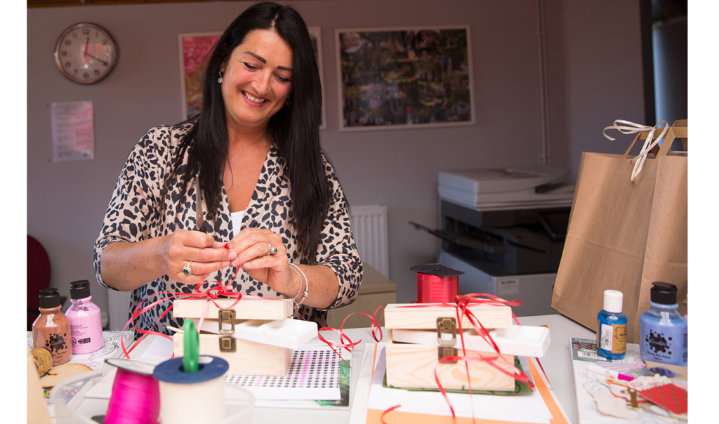 Adult white woman standing at a table with arts and crafts materials. She is smiling and tying a ribbon to a wooden box