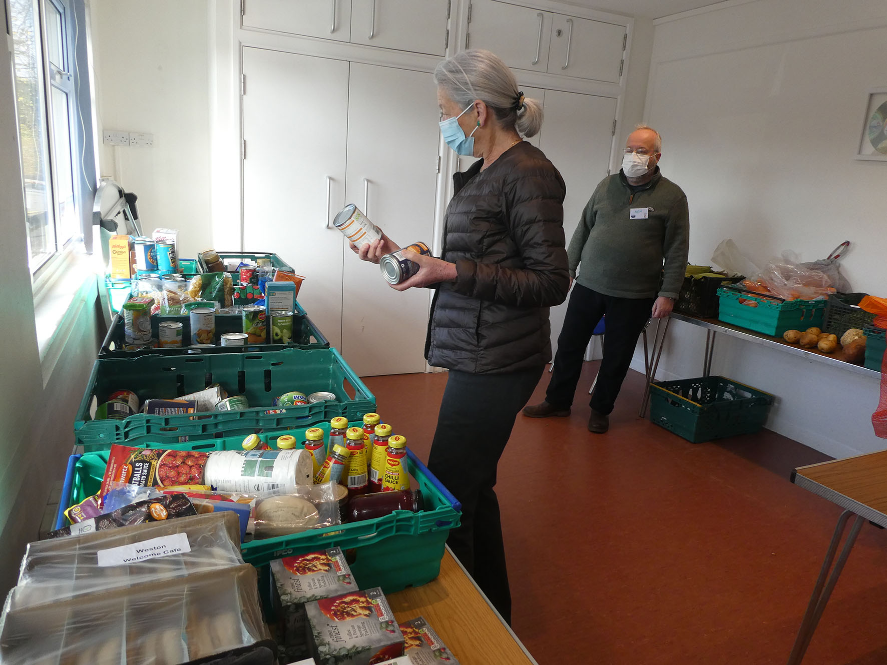 Elder white woman and elder white man wearing disposable medical masks. She carries two tins to a long table covered in large green plastic crates. In the crates is food, some tins, some jars, bottles and packets.ow. The