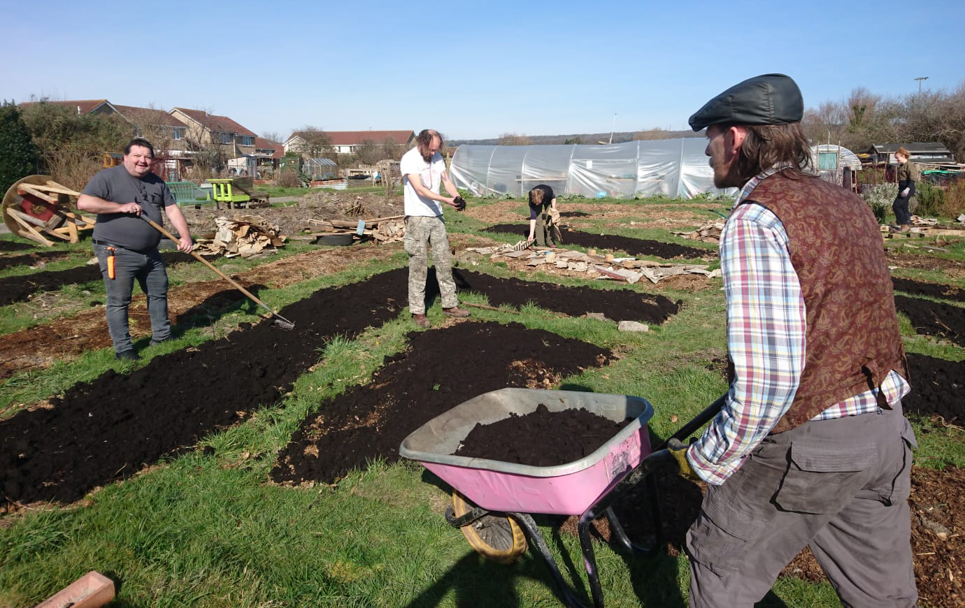 Six people work on the allotment. In the near foreground a man in flat cap pushes a wheelbarrow full of soil, to the left a man with a rake smiles to camera, in the mid foreground a man breaks up soil with his hands, in the mid distance a woman lays out equipment, while in the far distance a woman walks into the allotment