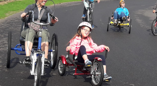 A learning disabled child on a tricycle on a outdoor track with others - she is wearing a pink coat and crash helmet. She has a look of joy and concentration as she squints into the sun. To her left is a smiling adult carer, also on a tricycle