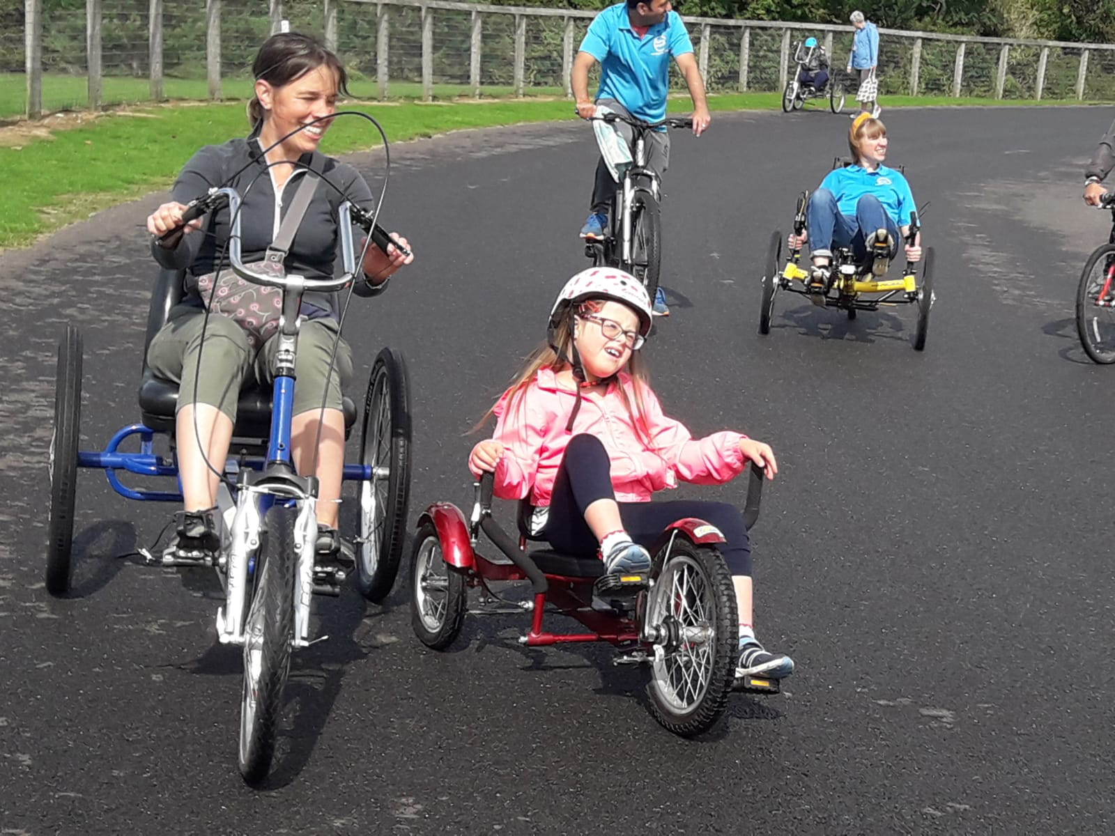 A learning disabled child on a tricycle on a outdoor track with others - she is wearing a pink coat and crash helmet. She has a look of joy and concentration as she squints into the sun. To her left is a smiling adult carer, also on a tricycle
