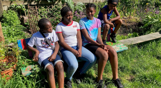 Four black school age children sit on a wooden bend outside