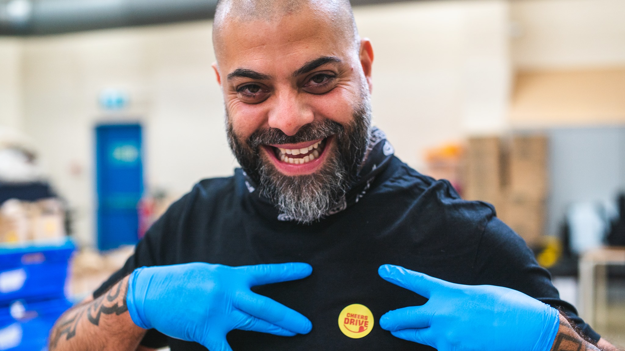 A smiling man gesturing towards a Cheers Drive badge on his blue chef's overalls