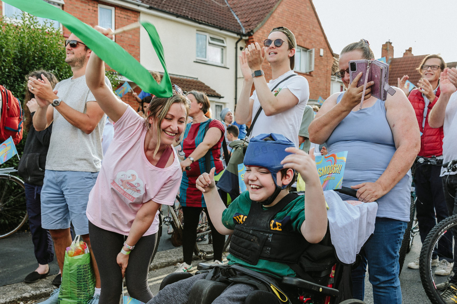 In the foreground a child in wheelchair beaming with joy with arms stretched up by his head dancing. To his left a laughing woman waves a green flag. The child's carer looks on smiling