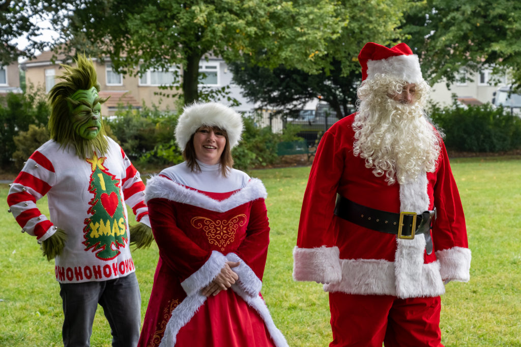 Three people in colourful costume. To the left a person dressed as the Dr Suess's The Grinch, in the middle a woman dressed as Mrs Claus and to the right, Santa with Hat and full white beard