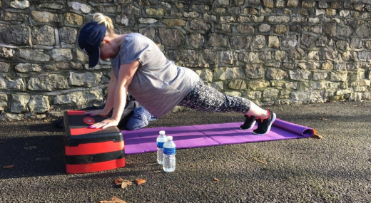 A person exercising outside in the sun on a colourful mat by a wall. They are performing 'plank'