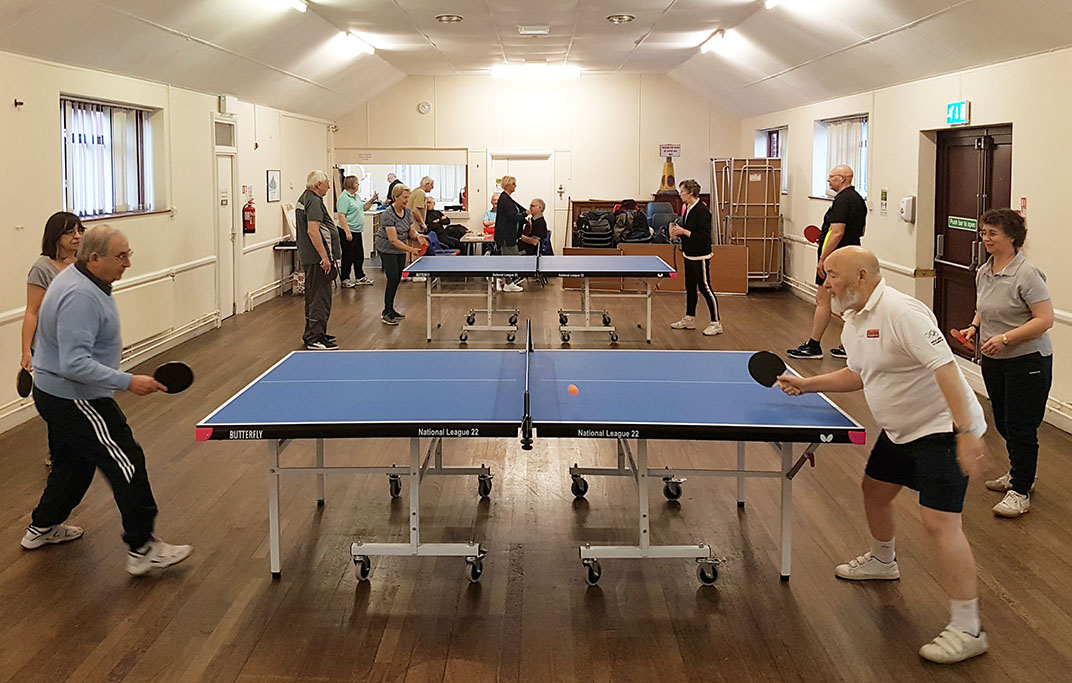 Elders in sports kit playing table tennis at two tables in a long hall